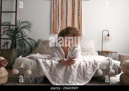 Smiling young lady lying on bed and using smartphone in morning Stock Photo
