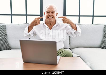 Senior man using laptop at home sitting on the sofa smiling cheerful showing and pointing with fingers teeth and mouth. dental health concept. Stock Photo