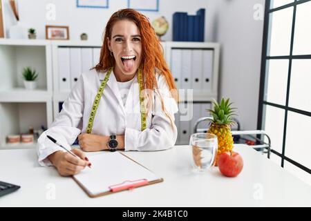 Young redhead woman nutritionist doctor at the clinic sticking tongue out happy with funny expression. emotion concept. Stock Photo