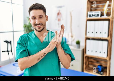 Young physiotherapist man working at pain recovery clinic clapping and applauding happy and joyful, smiling proud hands together Stock Photo