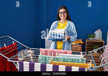 Young hispanic woman holding folded jeans hanging clothes on clothesline at laundry room Stock Photo