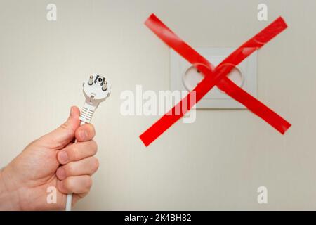A mans hand holds a white cord with a plug near an electrical outlet, tied with a red ribbon crosswise. Stock Photo