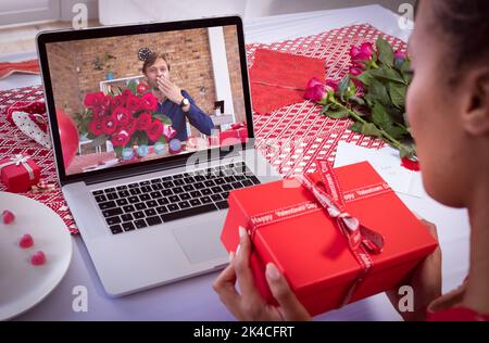 Diverse couple making valentines date video call on laptop holding gift and flowers. online valentines day during quarantine lockdown. Stock Photo