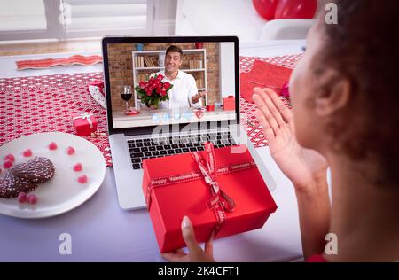 Diverse couple making valentines date video call on laptop holding gift and flowers. online valentines day during quarantine lockdown. Stock Photo