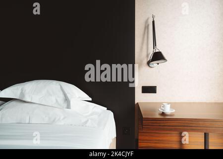 View of an empty hotel room with the made bed, black textured wall, night lights on the side over the desk with a cup of espresso coffee Stock Photo