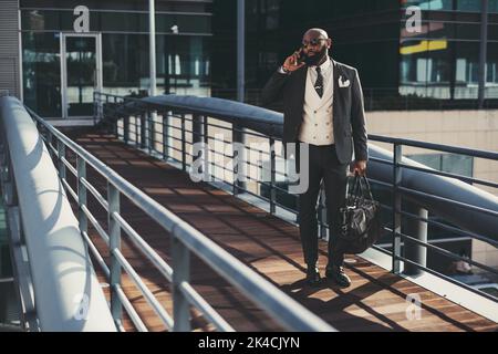 A mature fashionable black man entrepreneur is talking on the phone while passing on an overhead passage; an African bearded businessman is phoning wh Stock Photo