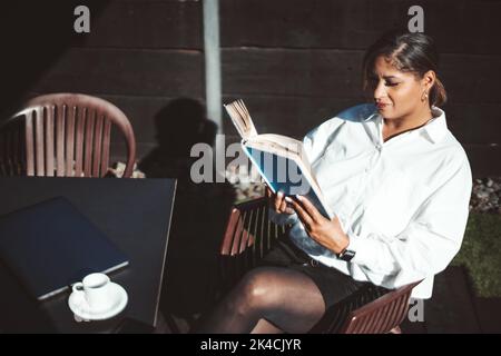 A portrait of a pensive mature plus-size Hispanic woman reading a book while sitting in an outdoor cafe on a warm autumn day with a cup of espresso co Stock Photo
