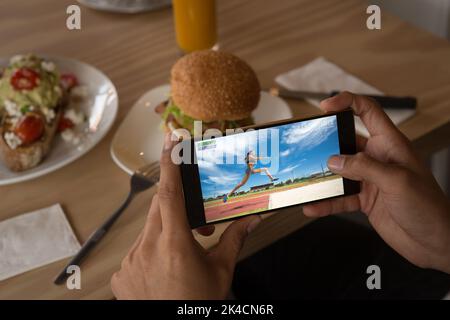 Hands of caucasian man at restaurant watching female athlete doing long jump on smartphone. sports, competition, entertainment and technology concept Stock Photo