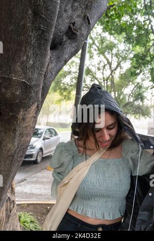 young woman putting on raincoat in a sudden unexpected rain, sheltering in a tree, wet, mexico Stock Photo