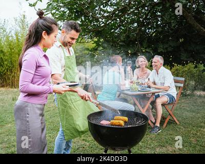 Couple cooking on a barbeque in countryside. At the back, his friends celebrating with alcohol drinks. Lifestyle concept. Stock Photo