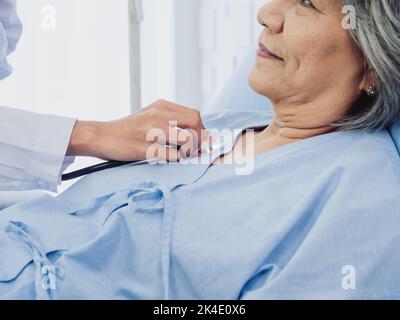 Closeup doctor's hand using stethoscope to examining, listen to the heartbeat of Asian elderly old female patient in light blue dress lying on bed in Stock Photo