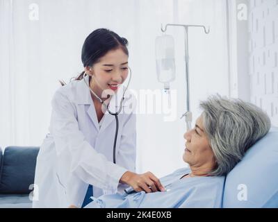 A beautiful young Asian woman doctor in white suit smiled while using stethoscope to examine, listen to heartbeat of elderly old female patient in blu Stock Photo