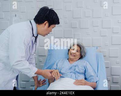 The kind Asian male doctor in white suit visits, talks and gives support, holding hand of happy elderly senior woman patient in light blue dress lying Stock Photo