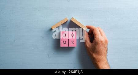 Male hand making a house made of four wooden blocks with letters on them to spell the word Home. Over pastel blue wooden background. Stock Photo