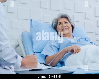 Happy and smiling Asian senior, old female patient lying on bed with receiving saline solution discussing with male doctor while he taking notes in ho Stock Photo