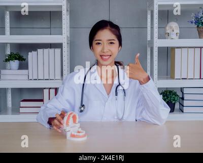 Young beautiful happy Asian female dentist in white suit thumbs up and smiles while holding and open jaw, dental tooth model on wood desk in office, l Stock Photo