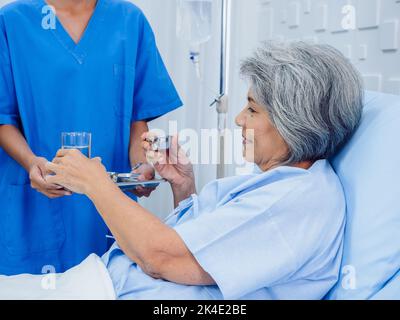 Asian elderly patient taking medicine, hold drinking water glass and pill from young nurse in blue scrub suit in hospital room, daily medicine or vita Stock Photo