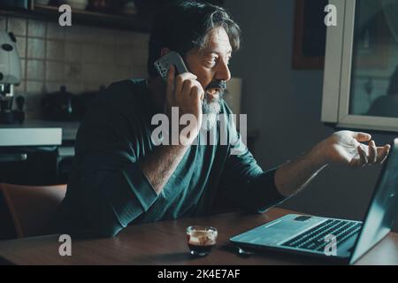 Mature young man working at home in the kitchen using laptop and calling with mobile phone. Concept of alternative workplace and smart working activit Stock Photo