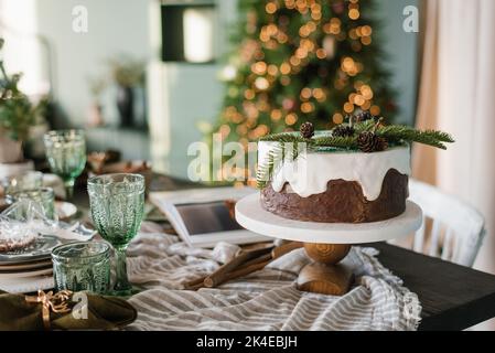Festive Christmas cake with white icing decorated with spruce branches stands on a set table against a background of lights Stock Photo