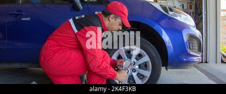 Picture of a mechanic in his workshop who with a pneumatic screwdriver disassembles the wheel of a car resting on trestles. Car repair work. Banner Stock Photo