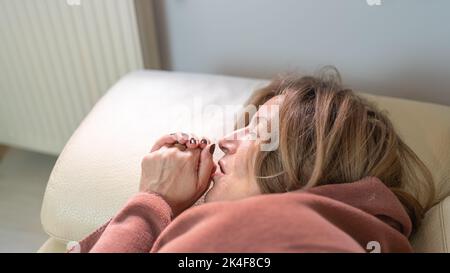 Woman lying on a sofa and going cold through the winter and energy saving, blowing her hands to warm herself. Stock Photo