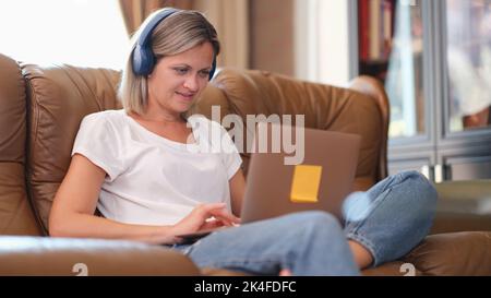 Smiling woman in headphones working on laptop on sofa Stock Photo