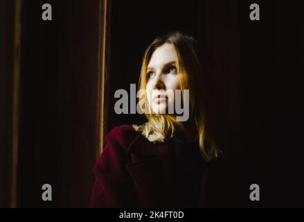 Portrait of a beautiful blonde woman in a burgundy coat, who is sitting alone and in the dark with a sad expression on her face. Stock Photo