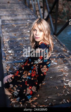 Calm female with blond hair wearing wearing dress and looking at camera while sitting on lumber bridge in fallen leaves Stock Photo