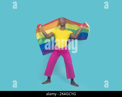 Full body barefoot African American man in colorful clothes with makeup covering head with LGBT flag against turquoise background Stock Photo