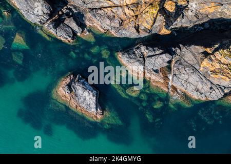 Aerial view directly above rugged rocky outcrop of land jutting into a, emerald green ocean with sand sea bed in Cornwall, UK with copy space Stock Photo