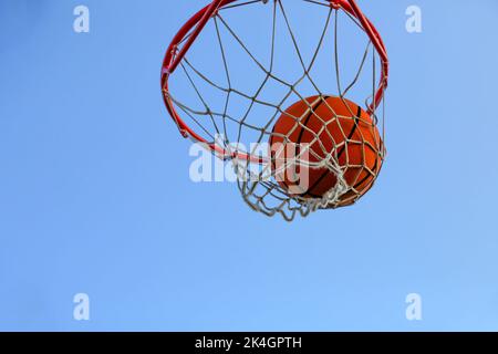 A basketball in a net against the sky. Sports, street game. The ball is in the ring. Hitting the target. Conceptual. Winning competitions. Success. Achieving the goal Stock Photo