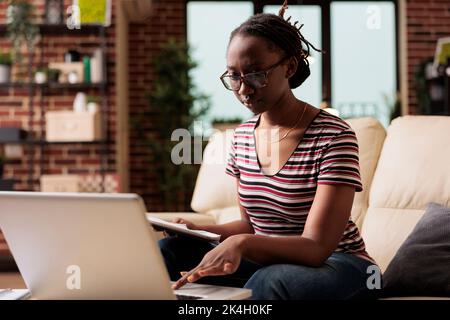Remote employee holding clipboard with report, working in home office, using laptop. Serious professional freelancer looking at computer, typing, busy remote employee in glasses Stock Photo