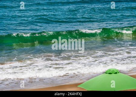 Green umbrella on the beach as the ocean waves come in with a big curl. Stock Photo