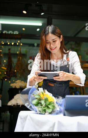 Portrait, Beautiful and pretty millennial Asian female florist or flower shop owner taking a picture of beautiful bouquet with her smartphone. Stock Photo