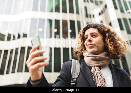 From below self assured mature male with curly hair reading message on cellphone while standing against modern building in downtown Stock Photo