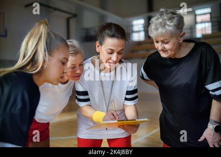 Female sport coach with clipboard discussing tactics with young and old women team training for match in gym. Stock Photo