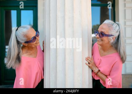 Senior women twins in colourful clothes in city, posing next to architectural column. Stock Photo