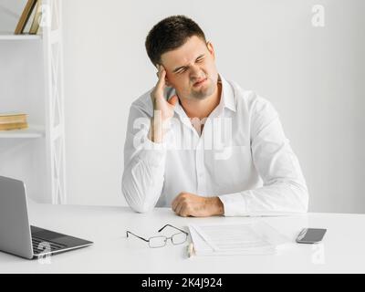 Exhausted work. Suffering man. Headache frustration. Sick guy holding head massaging temples taking over spectacles sitting desk light room interior. Stock Photo