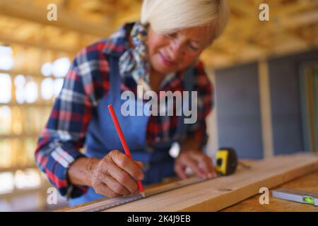 Senior woman measuring wooden board inside of her unfinished ecological wooden eco house. Stock Photo