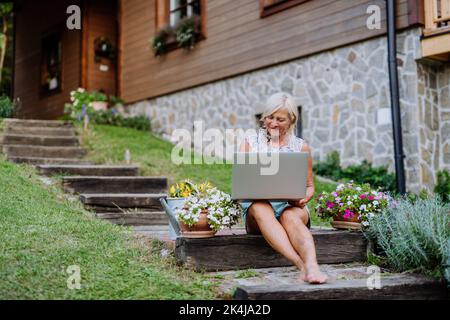 A senior woman using laptop and handling orders of her homegrown organic flowers and vegetables in garden Stock Photo