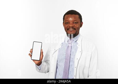 Smiling black doctor man with small beard in white coat holds smartphone blank mockup, isolated on white background. Happy black african american phys Stock Photo