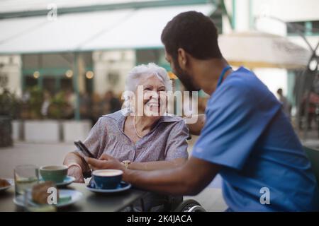 Caregiver having coffee with his client and learning her using a smartphone, outdoor at cafe. Stock Photo