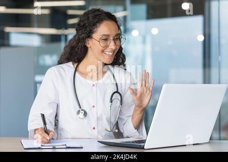The doctor is online. A young Latin American female doctor participates in an online medical conference, meeting. He waves his hand at the laptop camera, says hello, smiles, takes notes. Stock Photo