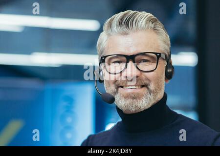 Portrait of a gray-haired handsome man in glasses and a headset with a microphone. Speaker, business trainer, online conference. Looking at the camera, smiling. Stock Photo
