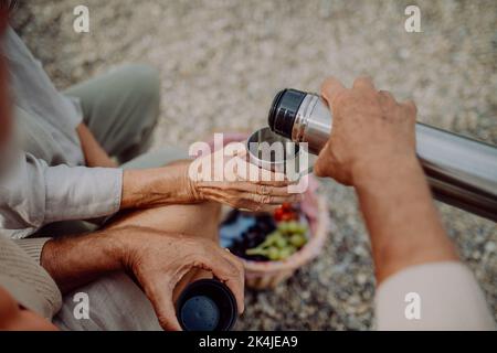 Close-up of senior couple having picnic at beach during autumn day, drinking hot tea from thermos. Stock Photo