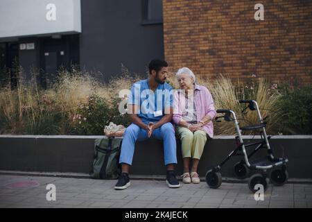 Happy senior woman sitting and talking with medical assistant in front of nurishing home. Stock Photo