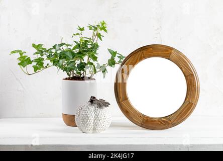 Empty round wooden frame, green houseplant ivy, ceramic pumpkin on white wooden table on textured wall backdrop. Modern home decor. Minimalistic conce Stock Photo