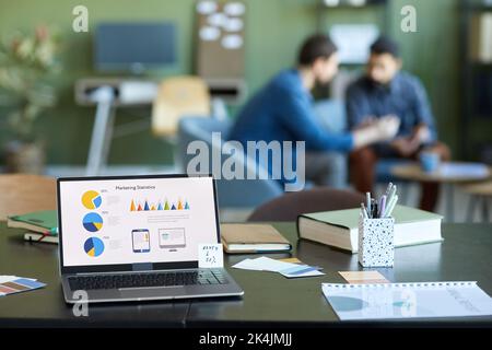 Laptop screen with financial data and variety of supplies of office worker on desk with two businessmen having discussion on background Stock Photo