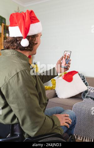 Caucasian man in wheelchair and santa hat making smartphone christmas video call with senior parents. christmas, festivity and communication technolog Stock Photo