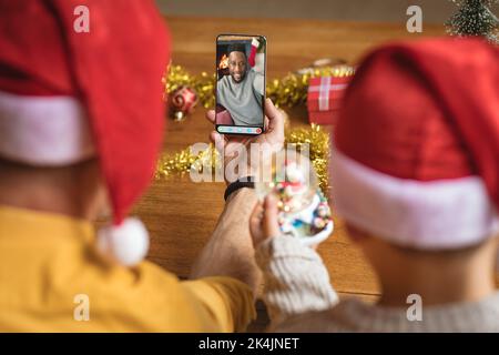 Father and son in santa hats making smartphone christmas video call with african american man. christmas, festivity and communication technology. Stock Photo